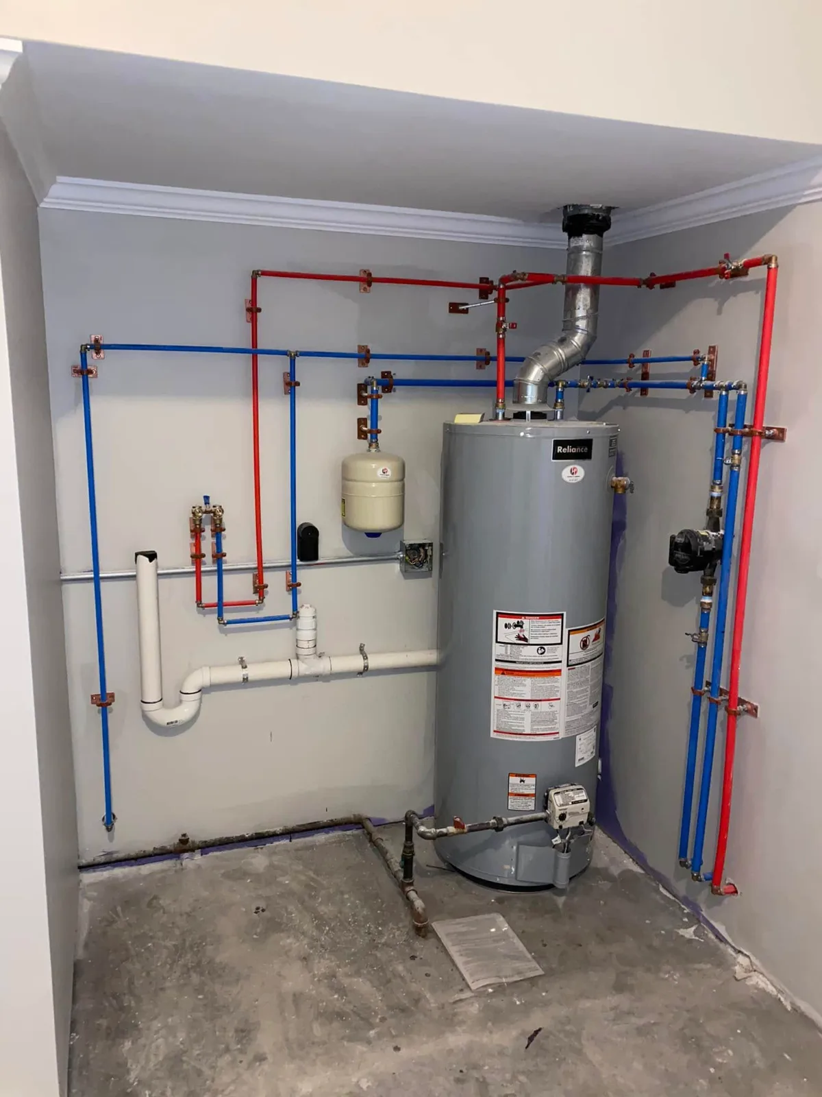 A gas water heater in a finished utility alcove, with red and blue water lines strapped along the walls, an expansion tank and a drain line