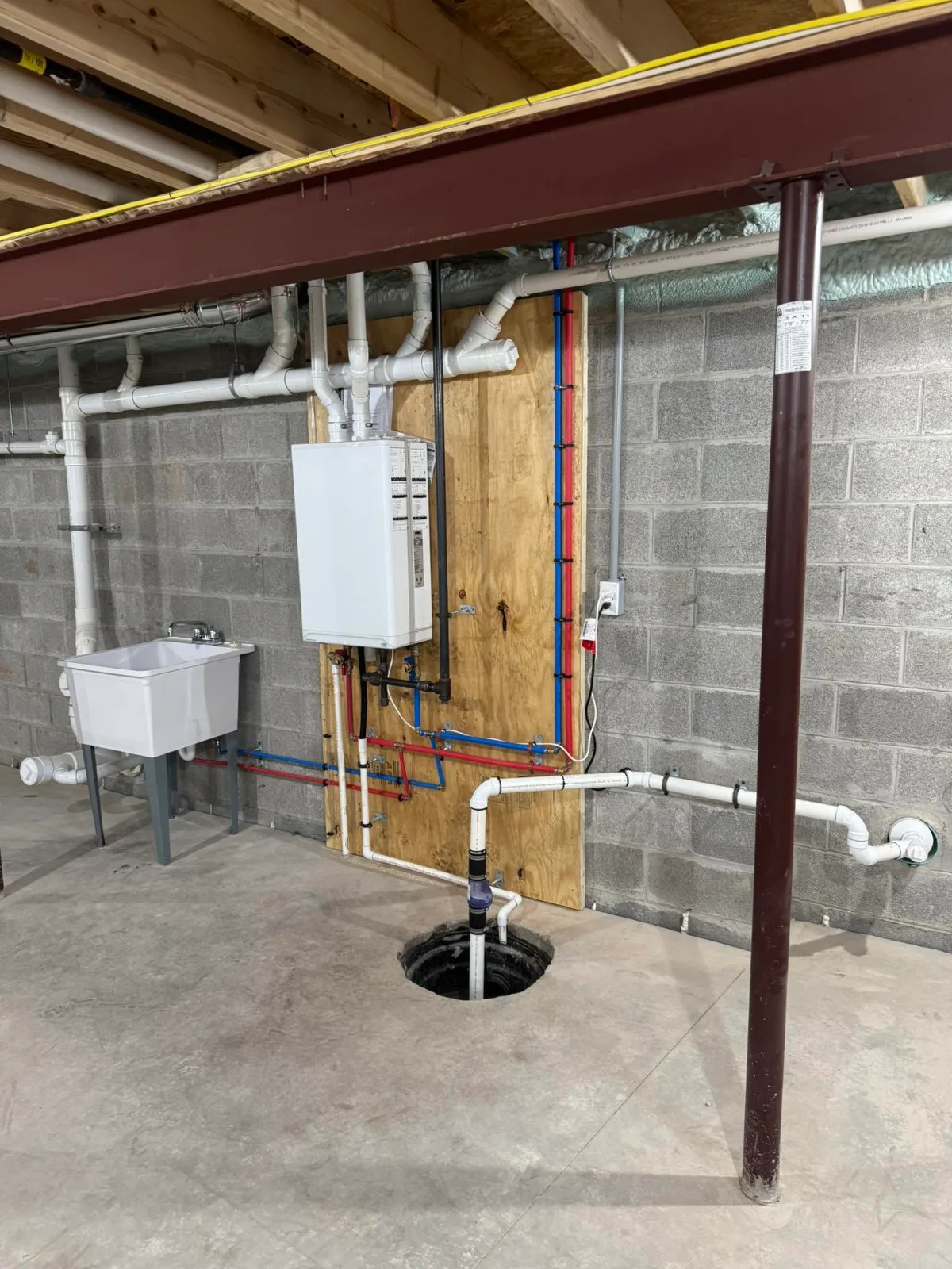 A finished basement utility area: a tankless water heater on a wood backboard, red and blue water lines, white drain pipes, a laundry sink and a sump pit