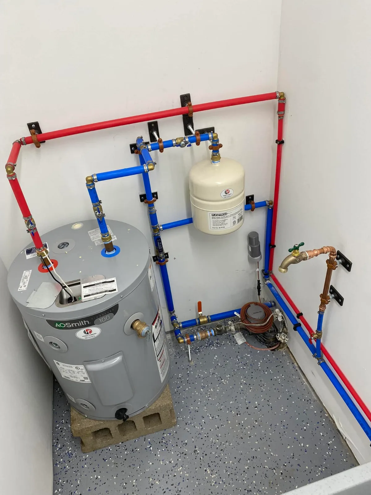 An electric water heater in a closet, with red and blue water lines, an expansion tank and a hose bib strapped to the wall