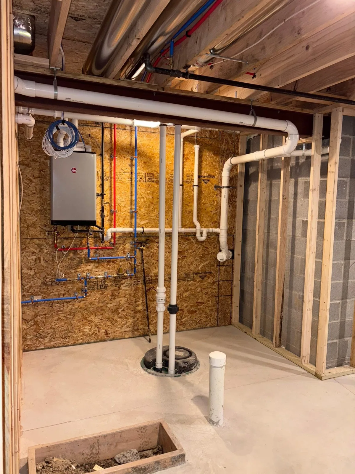 A basement mechanical room in progress: a Rheem tankless water heater, water lines, drain and vent piping, and the ejector basin in the floor