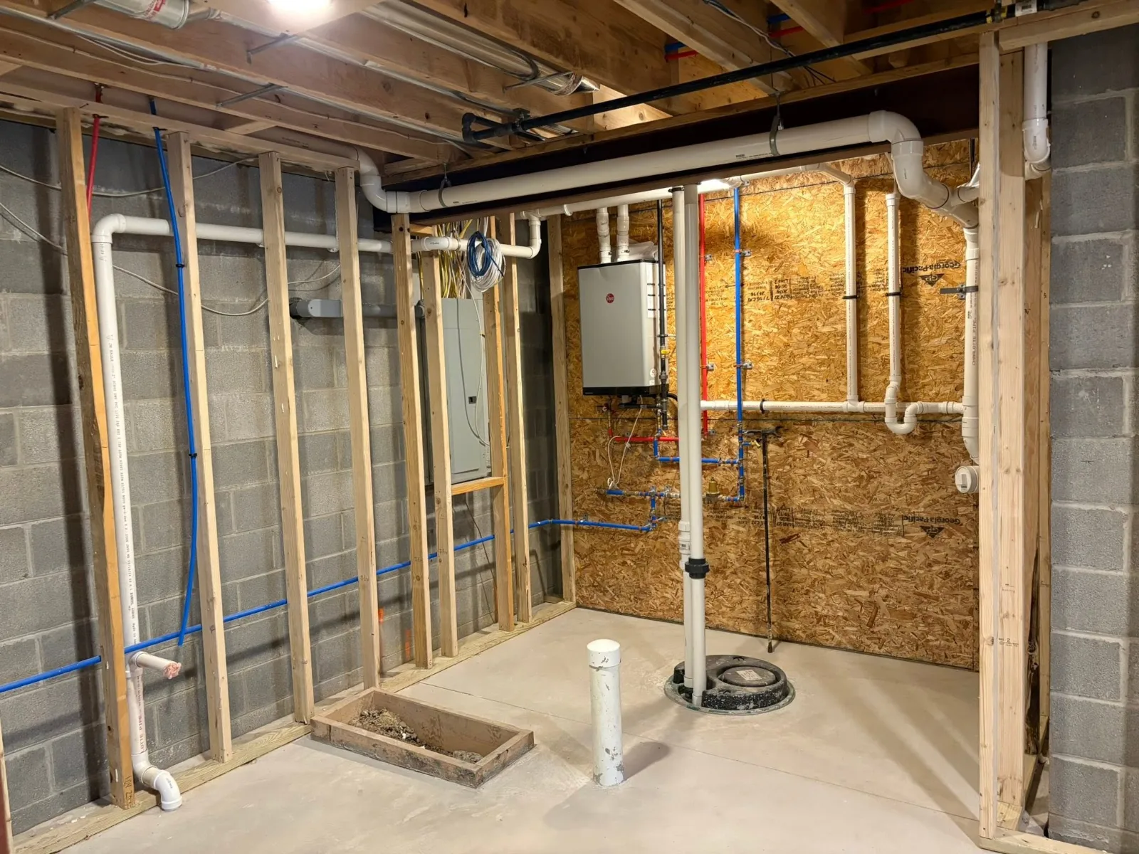 A basement mechanical room mid-build: a Rheem tankless water heater on a wood backboard, red and blue water lines, white drain and vent pipes, and a sewage ejector basin in the floor